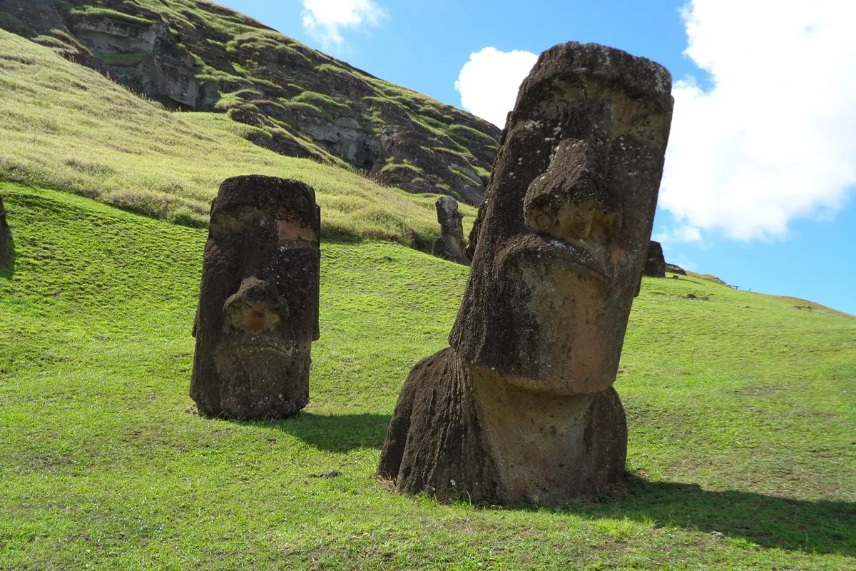 Moais da Ilha de Páscoa posicionados em uma encosta verde, sob céu azul com nuvens, representando a antiga cultura Rapa Nui.