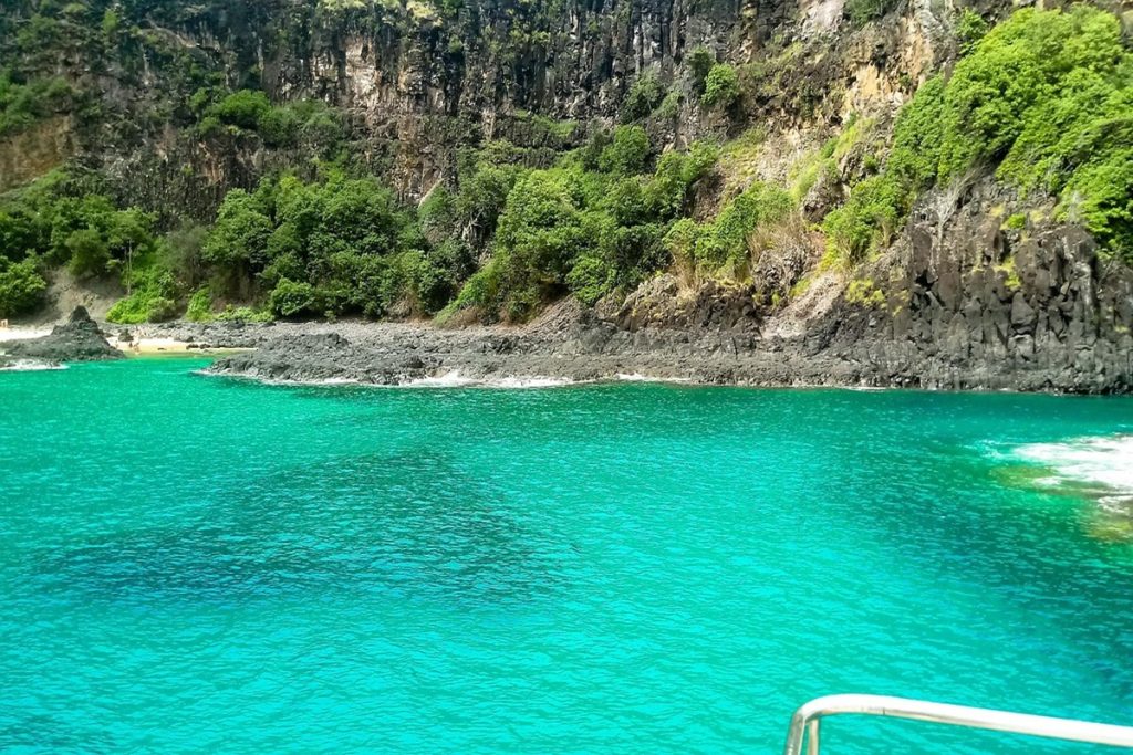 Mar cristalino em tons de azul-turquesa na Praia do Sancho, em Fernando de Noronha, com falésias cobertas por vegetação e formações rochosas.