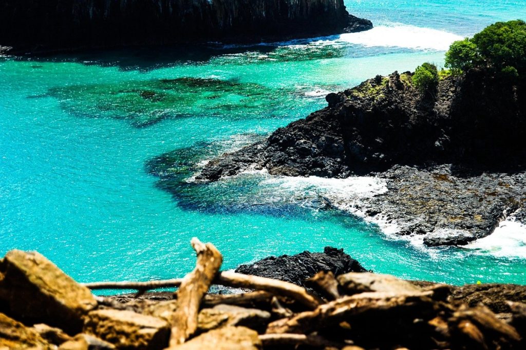 Vista aérea de uma enseada em Fernando de Noronha com mar azul-turquesa, pedras vulcânicas e vegetação tropical.