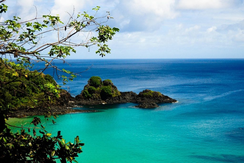 Vista panorâmica do mar azul-turquesa de Fernando de Noronha com formações rochosas e vegetação nativa.