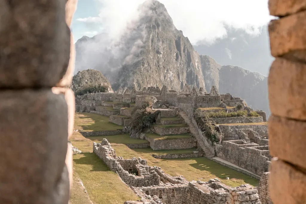 Onde ficar em Machu Picchu? Melhores opções de hospedagem Vista aérea das ruínas de Machu Picchu envoltas por névoa, com turistas caminhando entre os antigos muros de pedra e as montanhas verdes ao fundo.