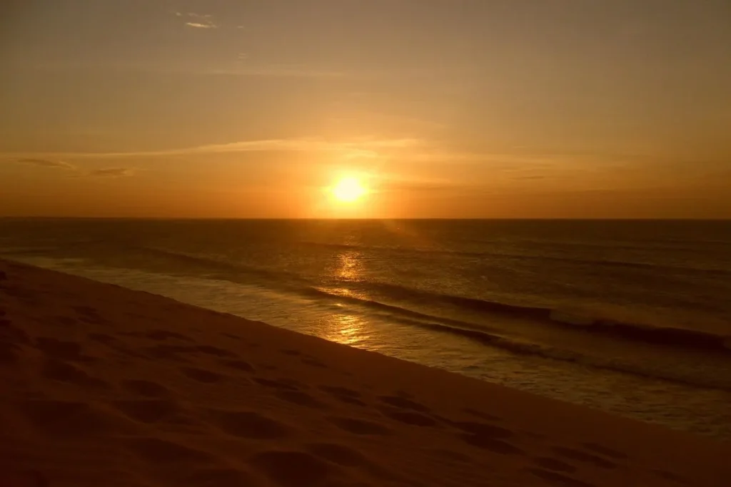Sol se pondo no mar visto das dunas de Jericoacoara, com céu alaranjado, ondas suaves e pegadas na areia.
