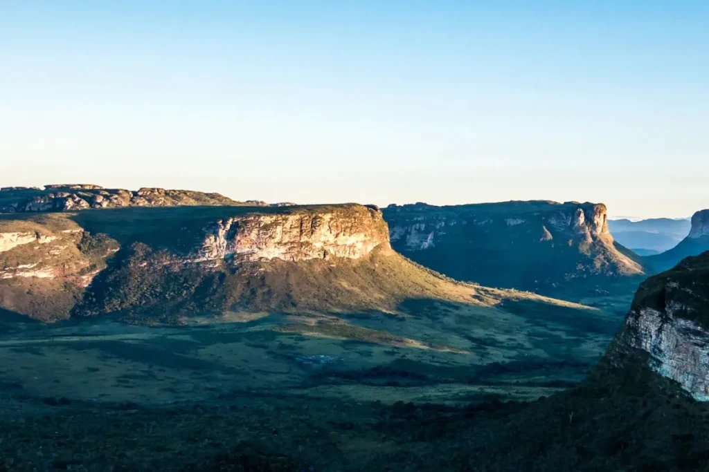 Vista panorâmica da Chapada Diamantina com paredões rochosos iluminados pelo sol, vegetação verde e céu azul claro.