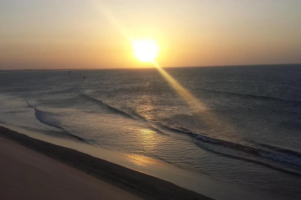 Sol se pondo sobre o mar em Jericoacoara, com reflexo dourado na água, pequenas ondas e duna à esquerda da imagem.