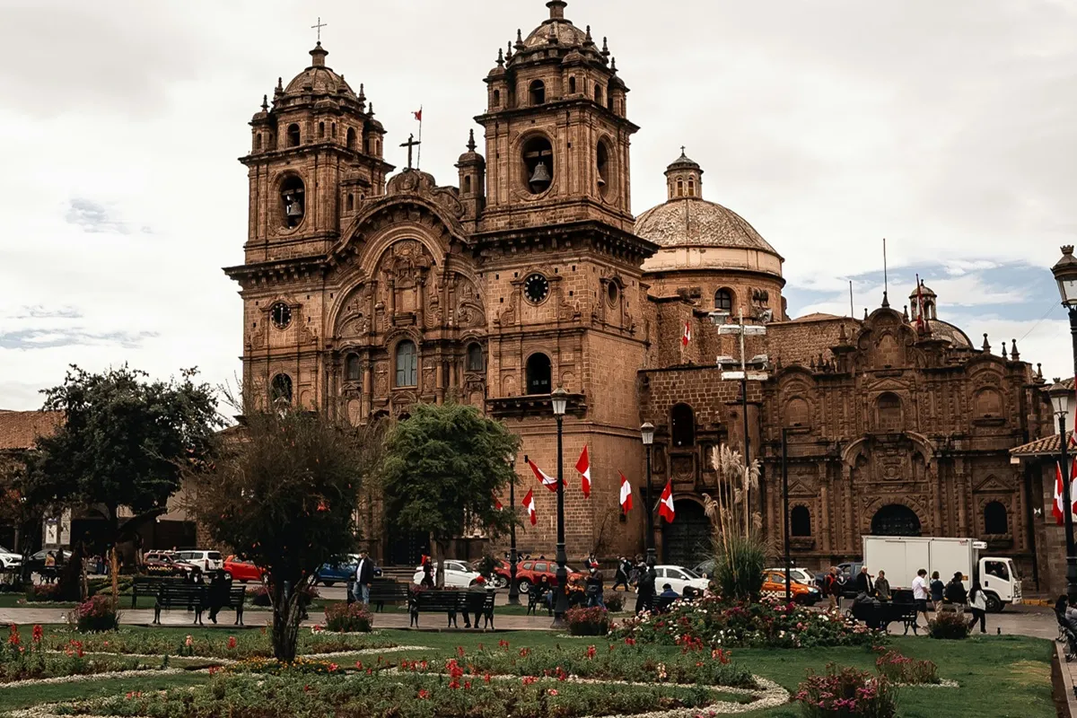 Vista frontal da Igreja da Companhia de Jesus em Cusco, Peru, com fachada barroca e bandeiras peruanas ao redor da praça central.