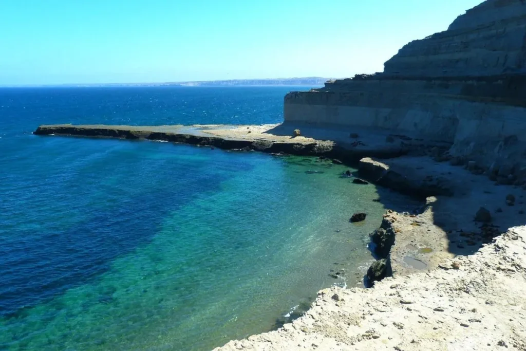 Vista panorâmica da costa de Puerto Madryn, na Patagônia, Argentina. A imagem mostra falésias rochosas íngremes e um mar calmo, com a água variando de um azul profundo a um turquesa claro próximo à praia.