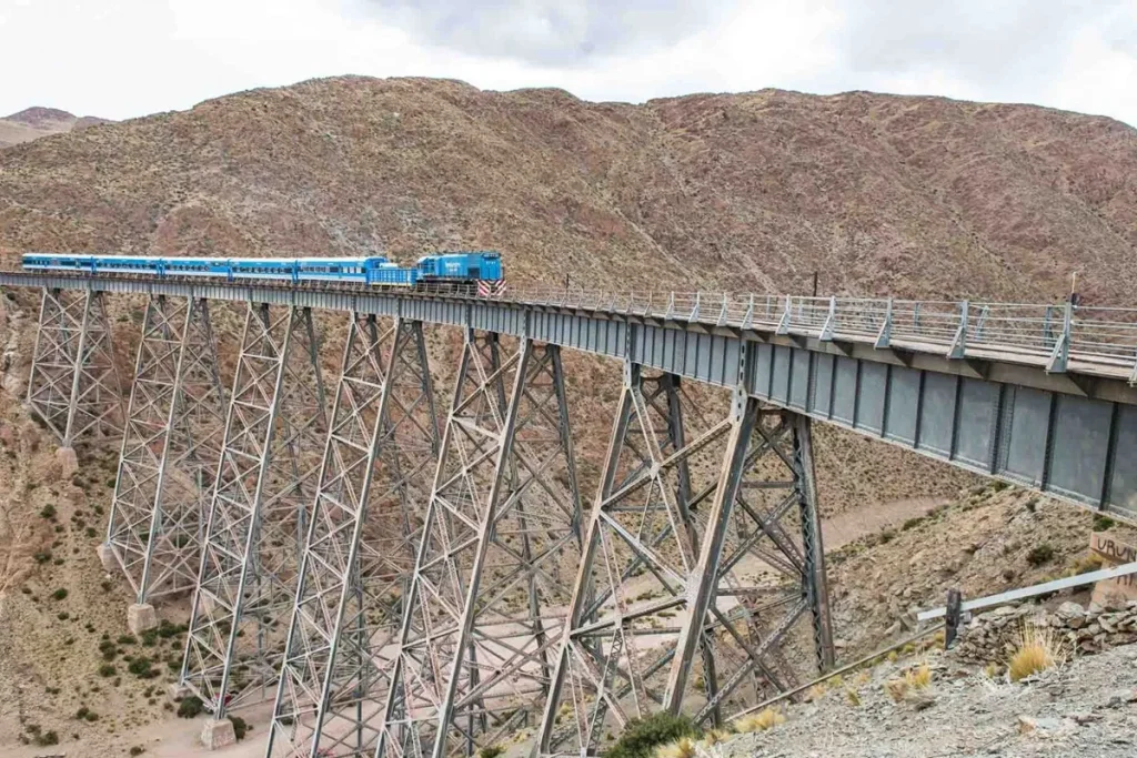Imagem do Trem das Nuvens atravessando uma ponte de ferro sobre um desfiladeiro, com paisagem de montanhas áridas ao fundo.