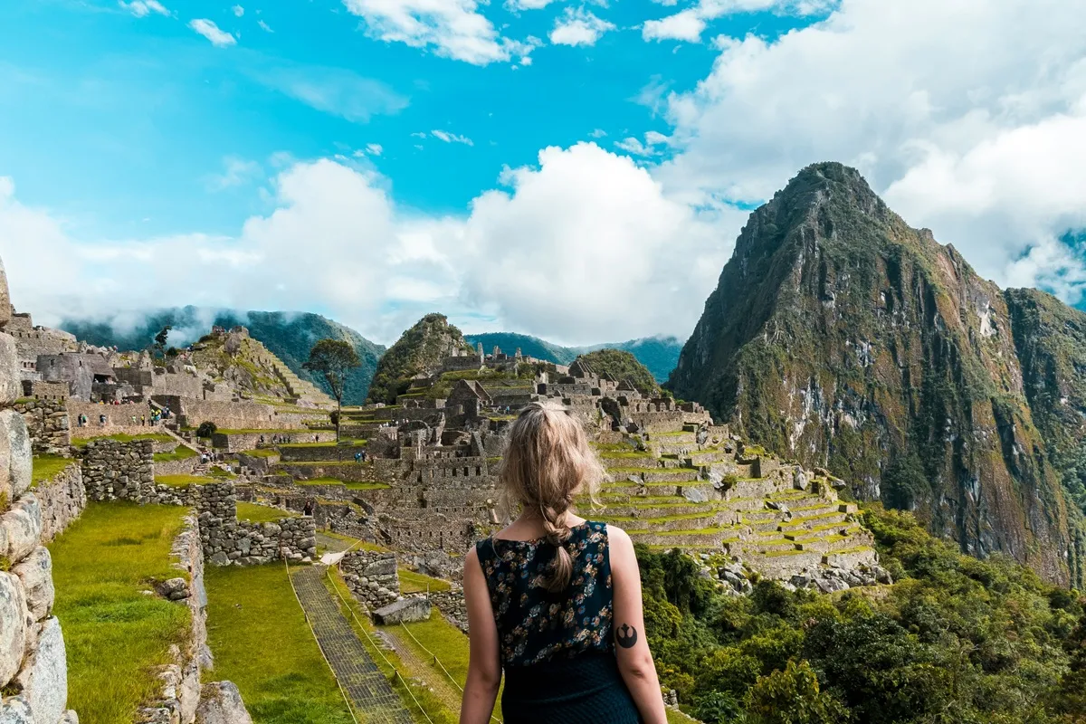 Mulher observando as ruínas de Machu Picchu com céu azul e montanhas ao fundo, em cenário típico dos Andes peruanos.