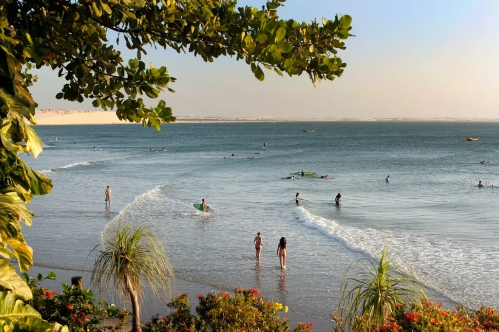 Praia de Jericoacoara ao entardecer, com banhistas, surfistas, coqueiros, vegetação e dunas ao fundo, no Ceará.