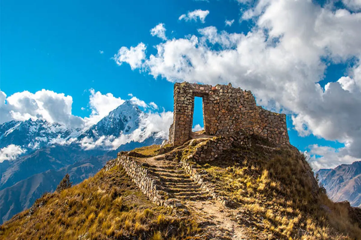 Ruína de portal de pedra em colina dos Andes com montanhas nevadas ao fundo sob céu azul com nuvens