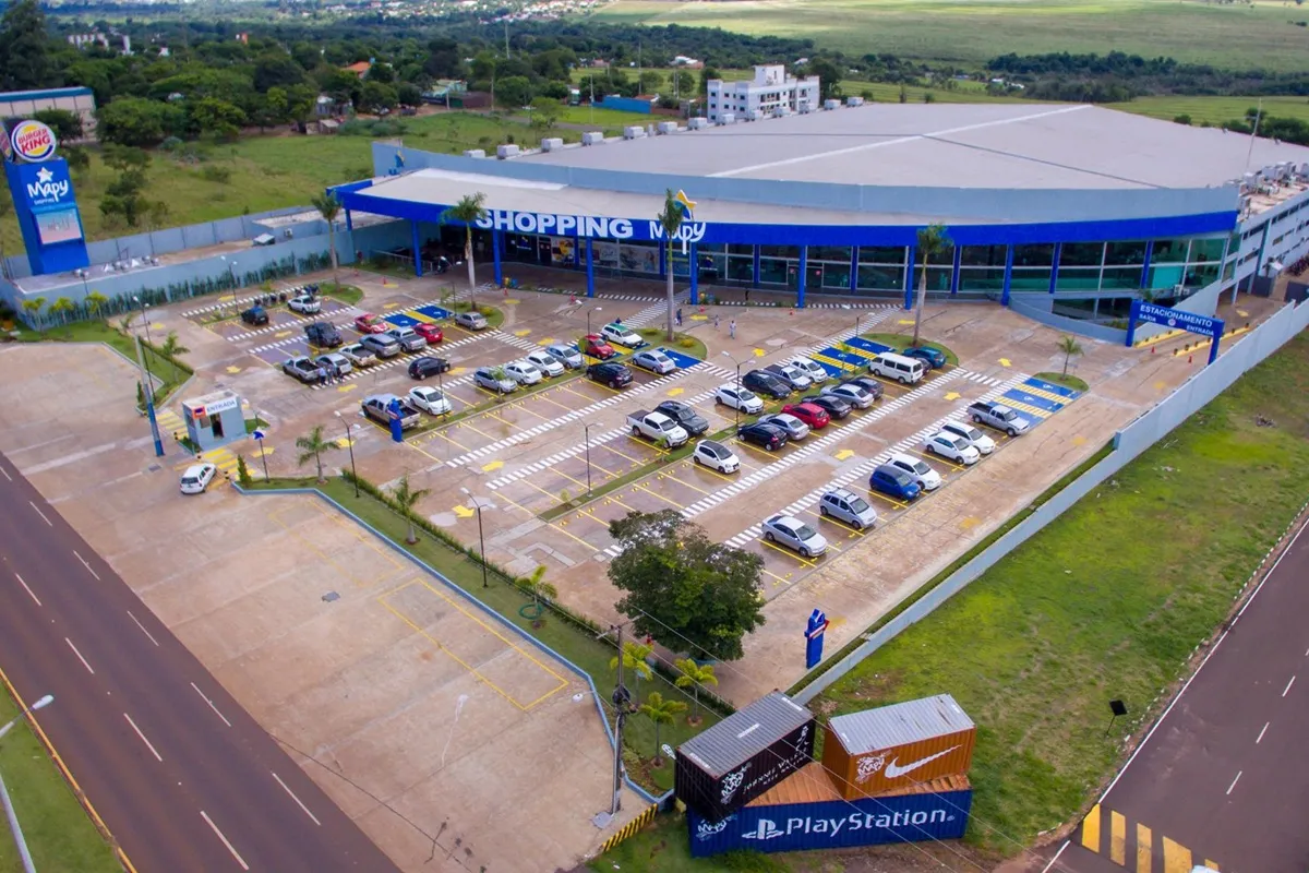 Vista aérea do Shopping Mbay em Salto del Guairá, com amplo estacionamento, veículos estacionados e contêineres de marcas como PlayStation em primeiro plano.