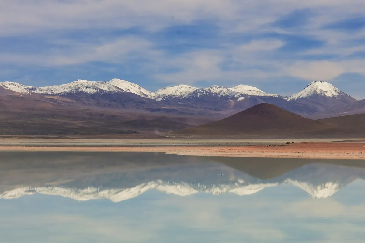 Vista panorâmica dos lagos andinos com montanhas cobertas de neve refletidas na água, no Deserto de Atacama