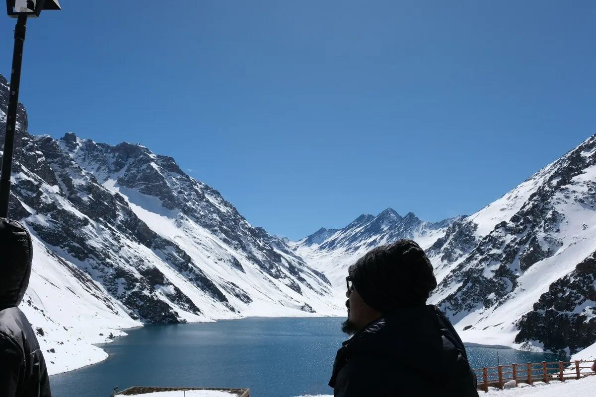 Turista observando lago cercado por montanhas nevadas sob céu azul na Cordilheira dos Andes no Chile
