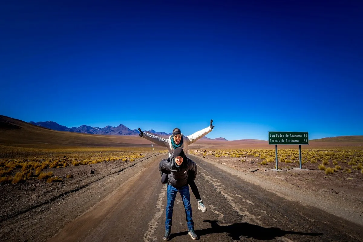 Duas pessoas se divertindo em uma estrada de terra no Deserto do Atacama, com um sinal de trânsito indicando "San Pedro de Atacama" e "Termas de Puritama", sob um céu azul, na região de Antofagasta.