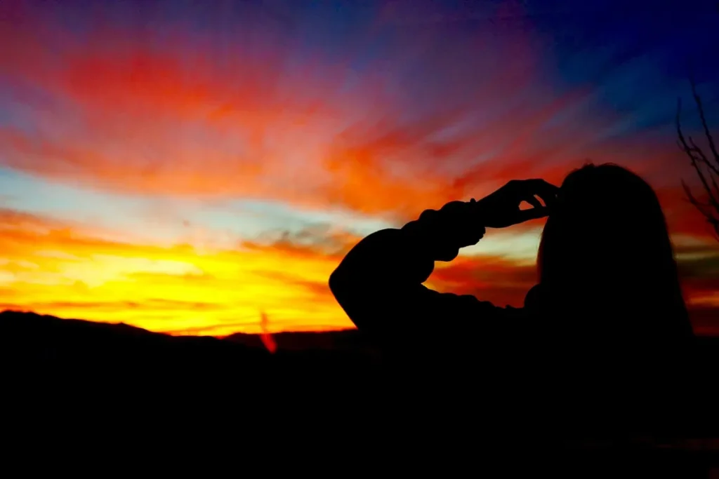 Pessoa em silhueta de perfil observando um pôr do sol alaranjado e púrpura sobre montanhas