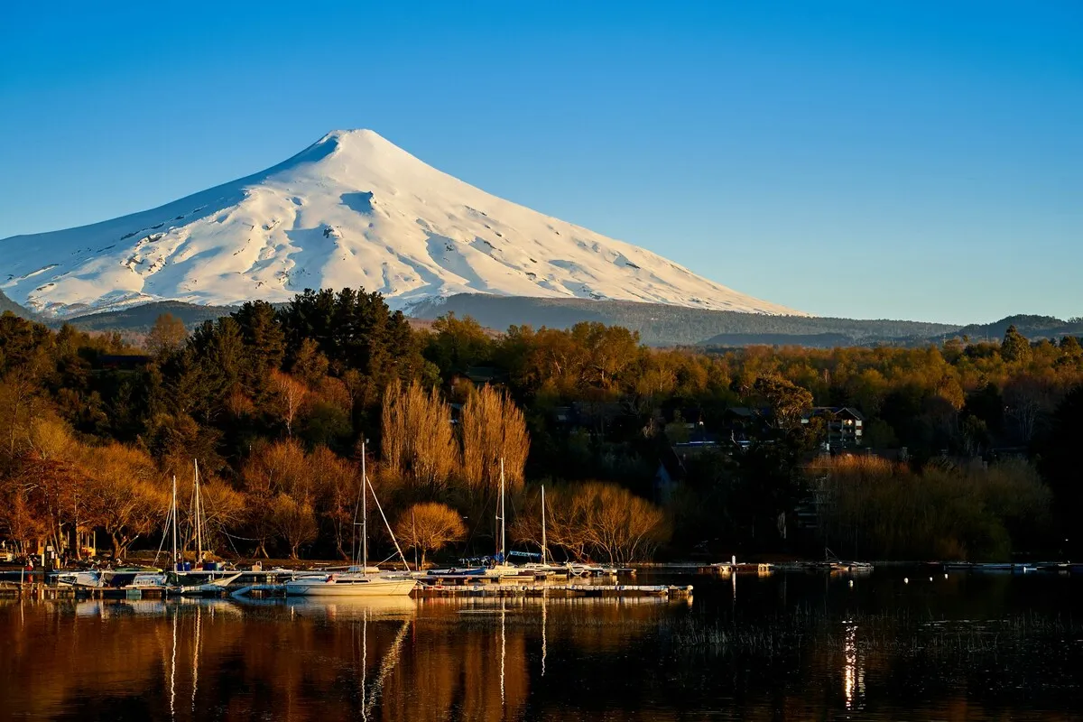 Vista panorâmica do Vulcão Villarrica coberto de neve, refletido nas águas calmas do Lago Villarrica, com barcos ancorados e árvores em tons de outono ou entardecer, mostrando a paisagem icônica de Pucón.