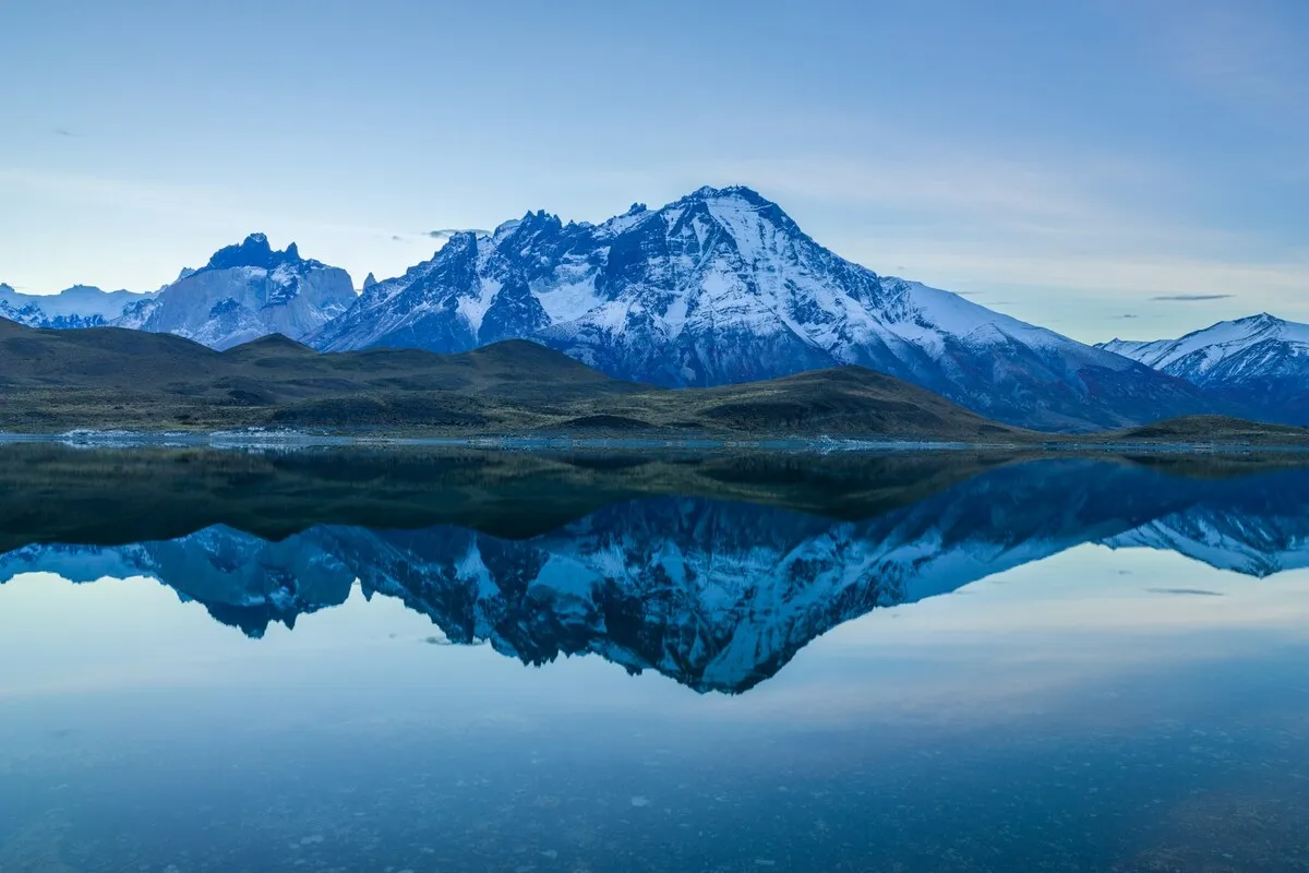 Imagem de montanhas nevadas refletidas em um lago calmo ao amanhecer ou entardecer, com a luz azulada típica de paisagens montanhosas, evocando a beleza de regiões como Valle Nevado e Farellones.