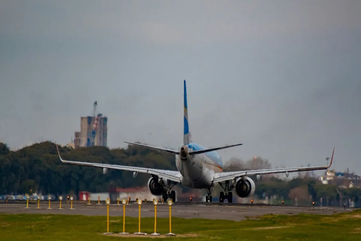 Vista traseira de um avião da Aerolíneas Argentinas acelerando na pista do Aeroporto de Bariloche, pronto para decolar, com luzes de pista em primeiro plano e edifícios e vegetação ao fundo, sob um céu nublado.