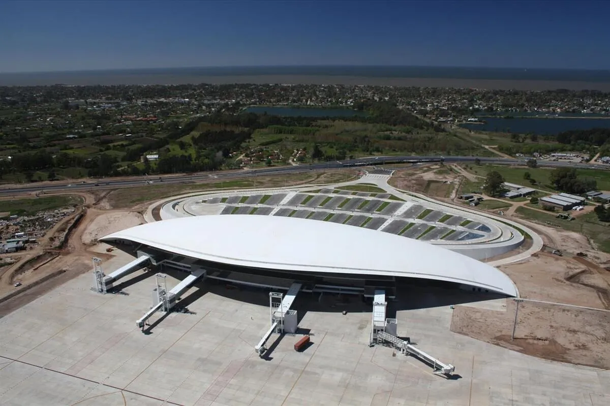 Vista aérea do Aeroporto Internacional de Carrasco, em Montevidéu, Uruguai, mostrando o terminal com seu distinto telhado curvo branco, pontes de embarque, pátio de aeronaves, estacionamento e a paisagem circundante com áreas verdes, estradas e corpos d'água ao fundo.
