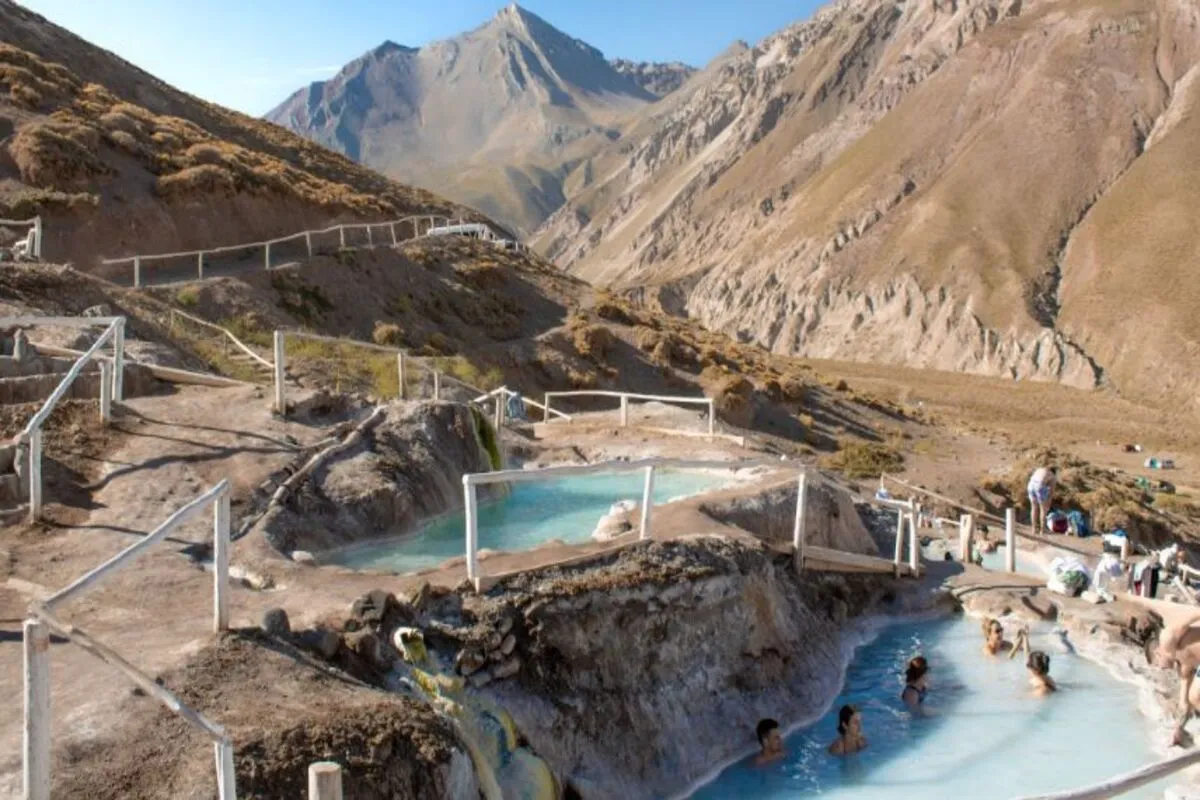 Piscinas naturais de água termal no Valle do Colina, Chile, com banhistas e paisagem montanhosa árida ao fundo.