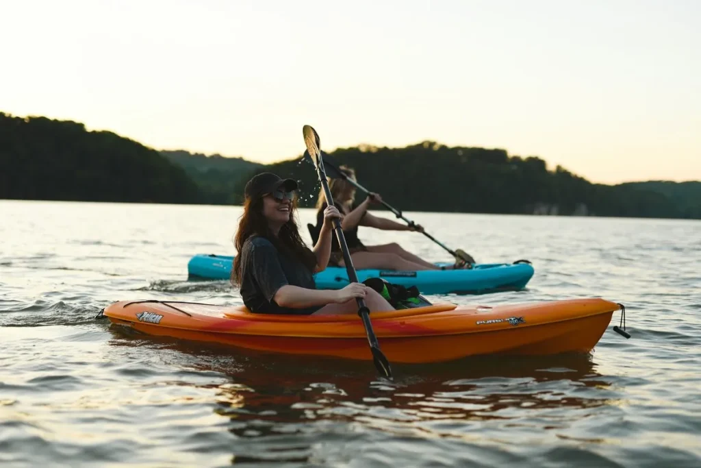 Duas mulheres em caiaques em lago calmo ao pôr do sol, desfrutando da natureza e do esporte.