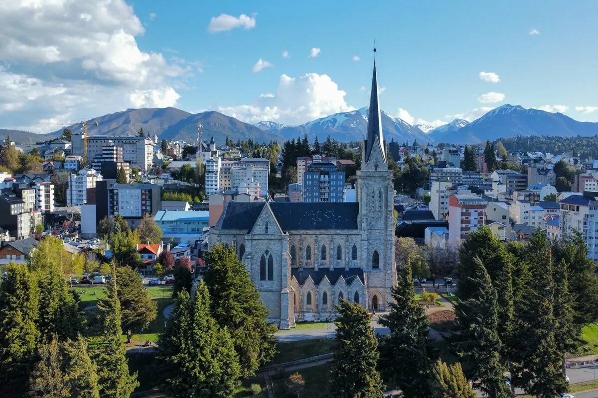 Vista aérea da Catedral de Bariloche com montanhas nevadas ao fundo, cidade de Bariloche em dia ensolarado de dezembro, paisagem de verão com picos brancos.