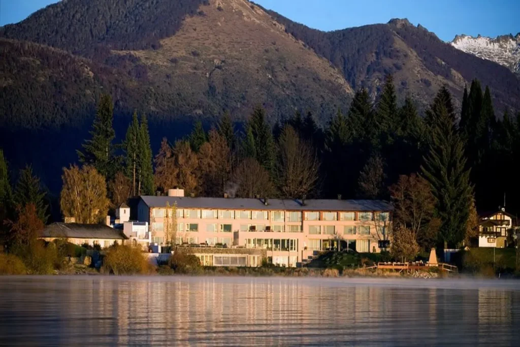 Grande hotel à beira do lago em Bariloche, rodeado por montanhas e florestas, com neblina sobre a água.