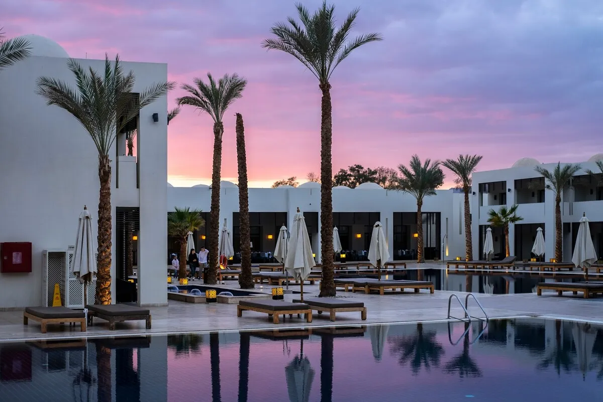 Vista noturna ou ao entardecer de um hotel de luxo com arquitetura branca e moderna. A grande piscina reflete o céu roxo e rosa de um pôr do sol vibrante, cercada por palmeiras altas, espreguiçadeiras e guarda-sóis fechados, criando um ambiente tranquilo e convidativo no hotel.