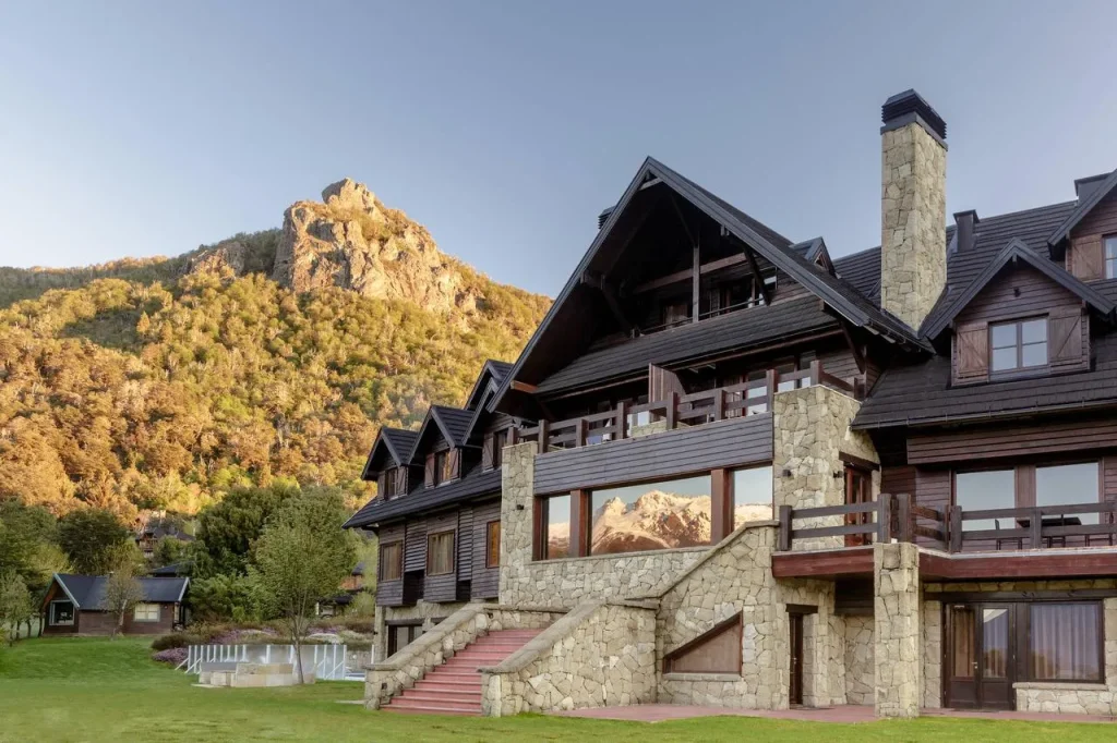 Fachada de hotel estilo chalé alpino em pedra e madeira, com varandas e janelas amplas, rodeado por gramado verde e montanha arborizada ao fundo