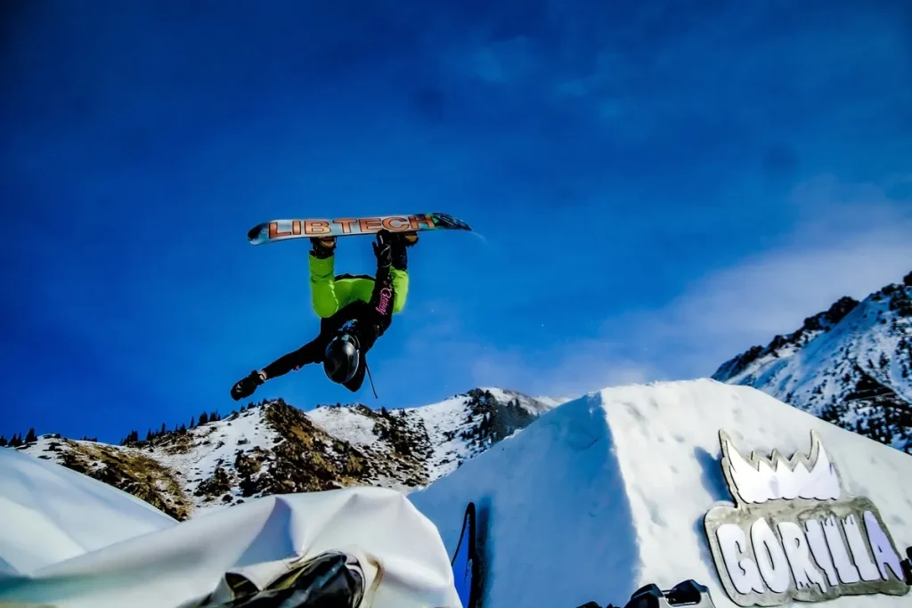 Snowboarder de cabeça para baixo segurando a prancha Lib Tech durante um salto sobre rampa de neve com o logo Gorilla ao fundo.
