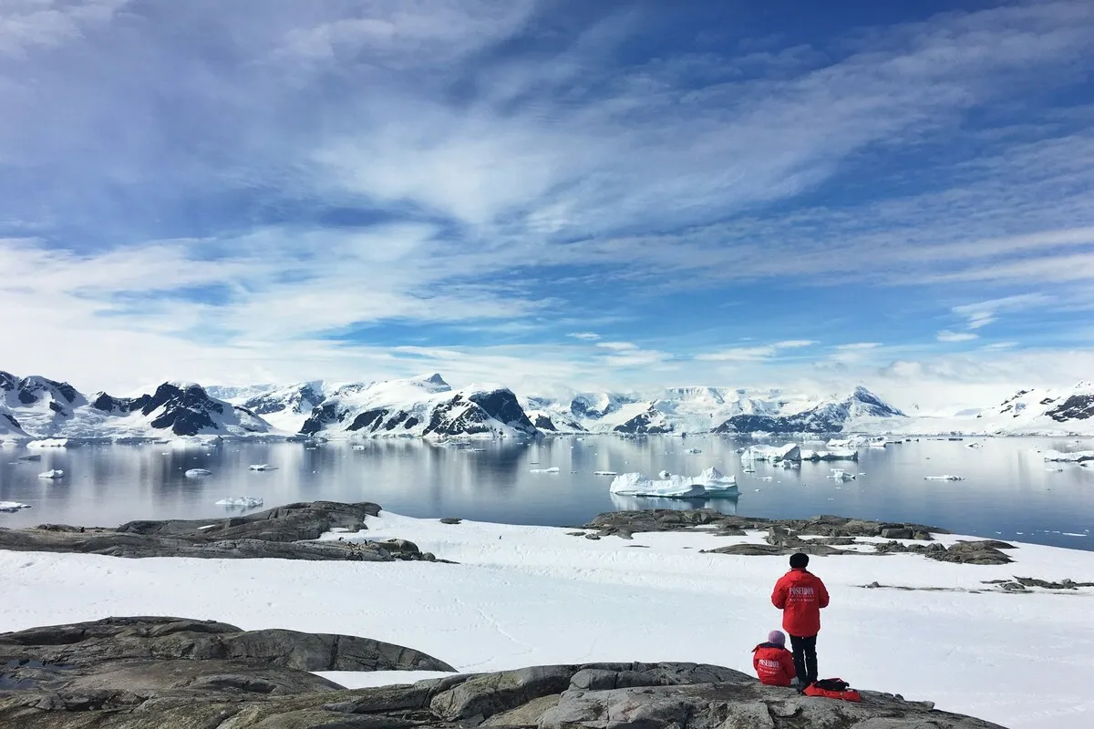 Duas pessoas, vestindo casacos vermelhos, em primeiro plano, observam a paisagem deslumbrante da Antártida, com montanhas nevadas e icebergs flutuando em águas calmas.