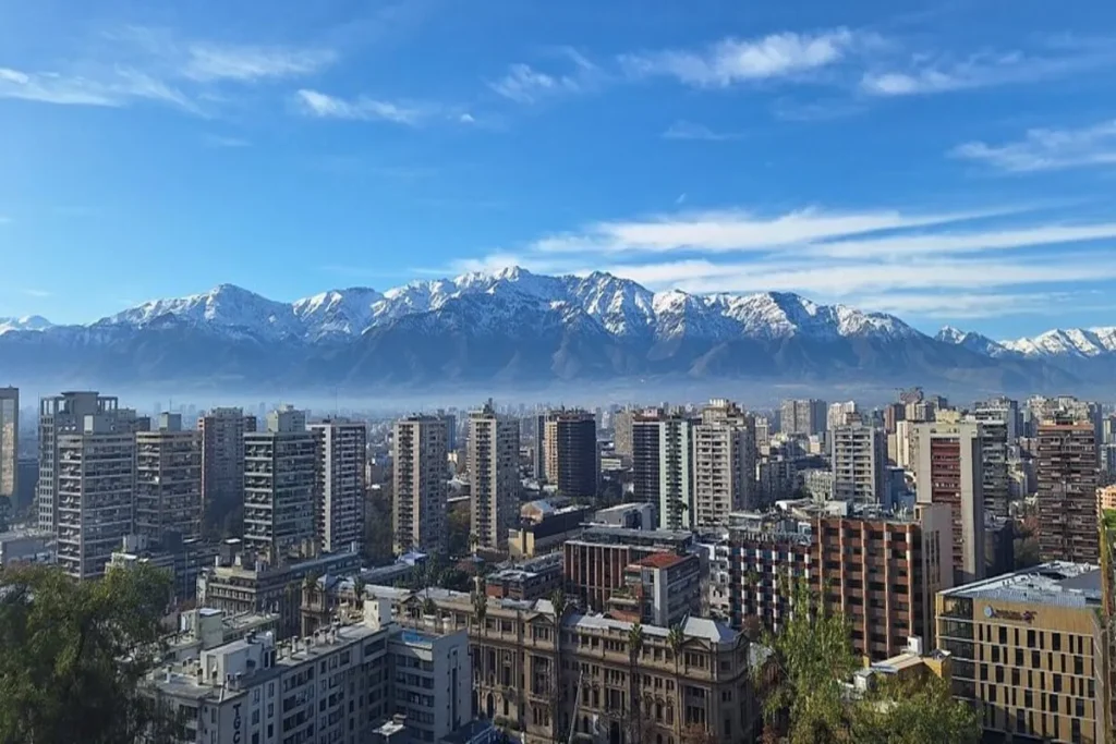 Vista panorâmica da paisagem urbana de Santiago a partir do Cerro Santa Lucía, com a Cordilheira dos Andes nevada ao fundo e o céu azul.