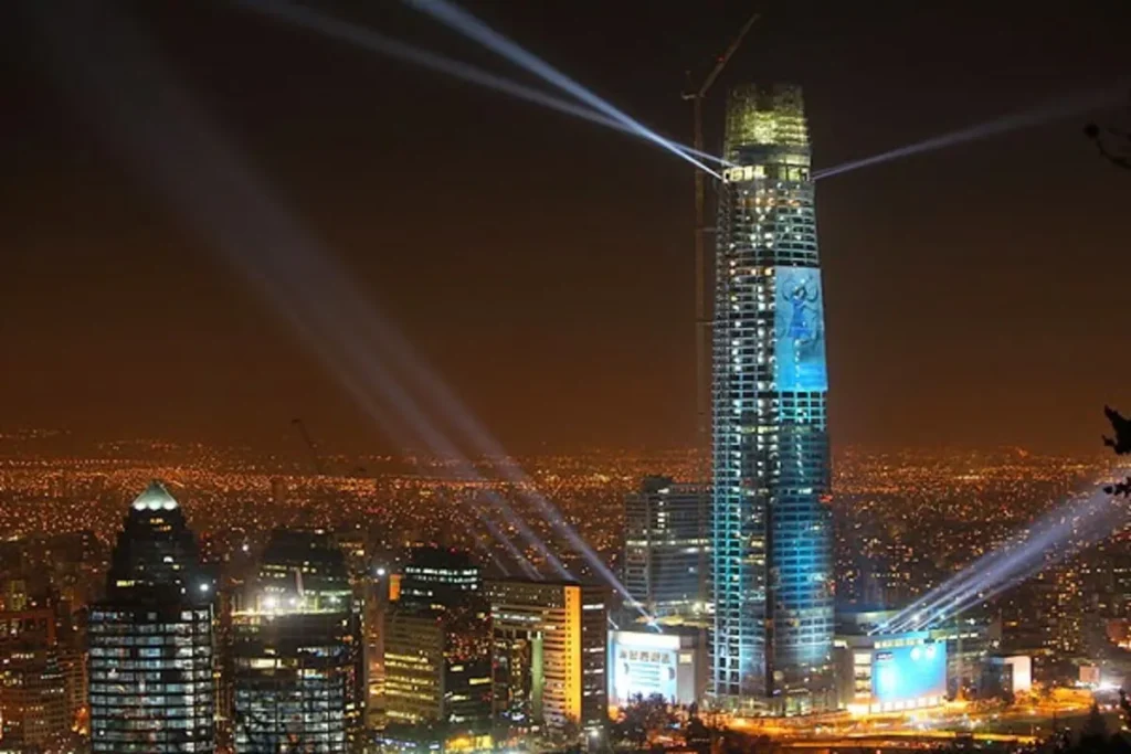 O arranha-céu iluminado do Costanera Center, com feixes de luz cruzando o céu noturno de Santiago, e a cidade abaixo, cheia de luzes.