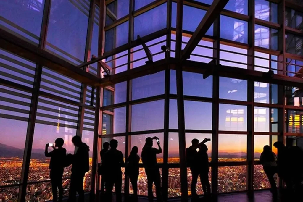 Silhuetas de pessoas observando o pôr do sol sobre a cidade de Santiago, Chile, de um deck de observação com janelas de vidro. A paisagem urbana está iluminada com luzes noturnas.