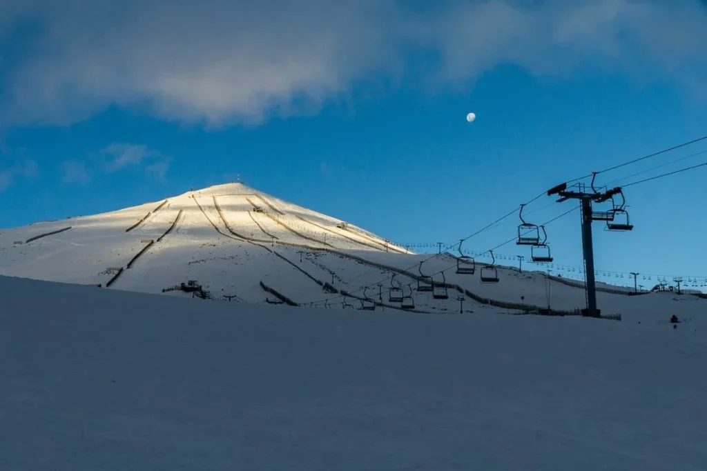 Pistas de esqui cobertas de neve na estação de El Colorado, com um teleférico em primeiro plano e a lua visível no céu azul.