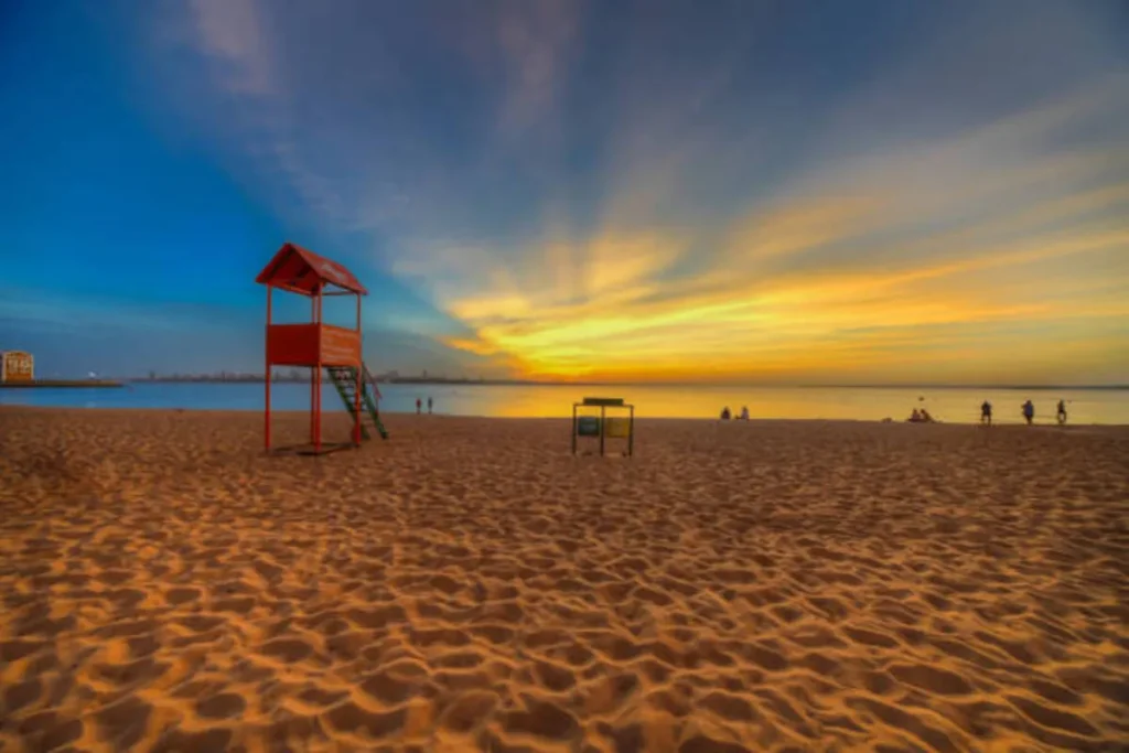 Pôr do sol dramático em praia de Encarnación, Paraguai, com céu em tons de azul e laranja, e posto de salva-vidas vermelho no primeiro plano.