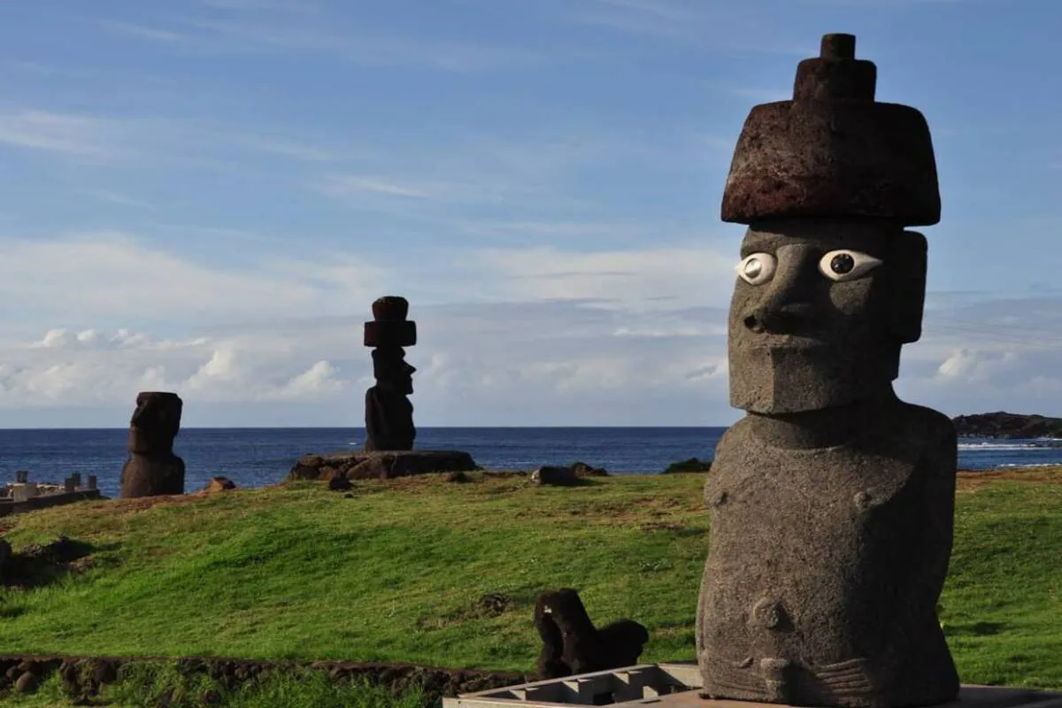 Estátuas Moai, algumas com pukao (chapéu) e olhos, em um campo gramado com o oceano ao fundo, na Ilha de Páscoa, próximo a Hanga Roa