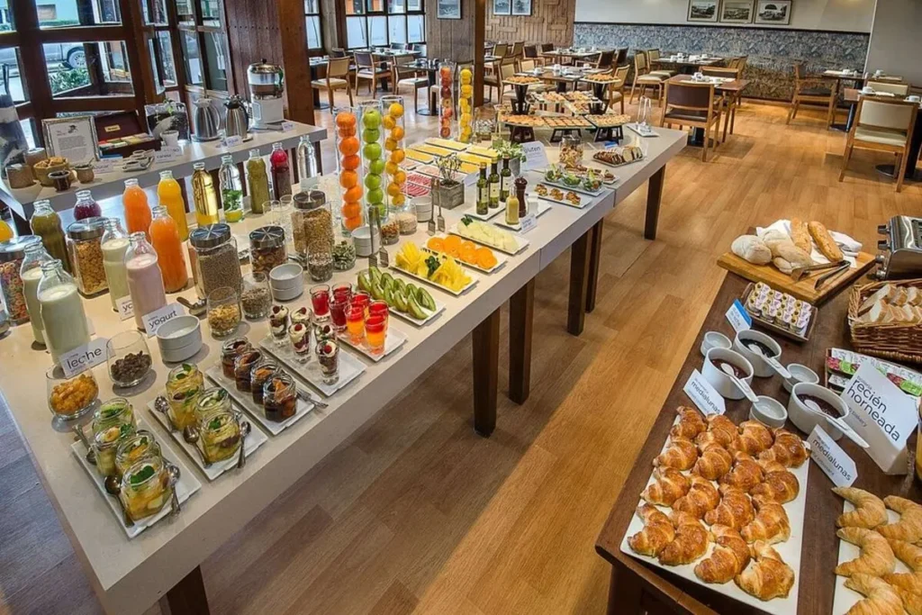 Grande buffet de café da manhã com uma variedade de alimentos e sucos, em um salão do Hotel NH Bariloche Edelweiss.