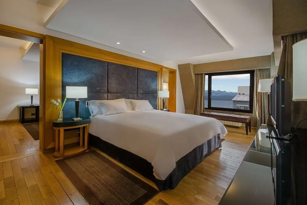 Quarto de luxo do Hotel NH Bariloche Edelweiss, com cama king-size, piso de madeira e vista panorâmica para o Lago Nahuel Huapi.