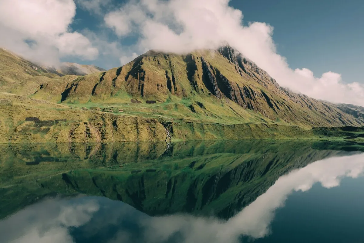 Paisagem em Huaraz, Peru, com um grande lago de água calma que reflete perfeitamente uma montanha verde e rochosa sob o céu nublado.