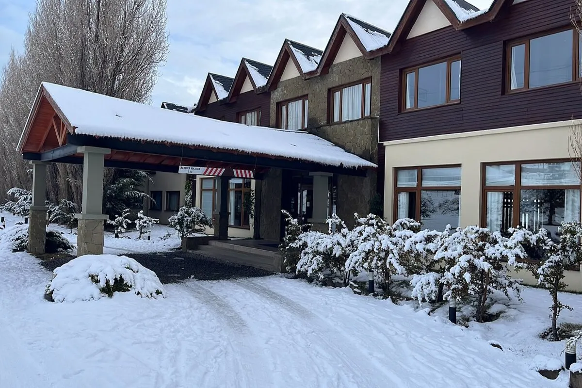 Exterior do Rochester Hotel El Calafate em um dia de inverno, com a fachada do hotel, o telhado e os arbustos cobertos de neve.