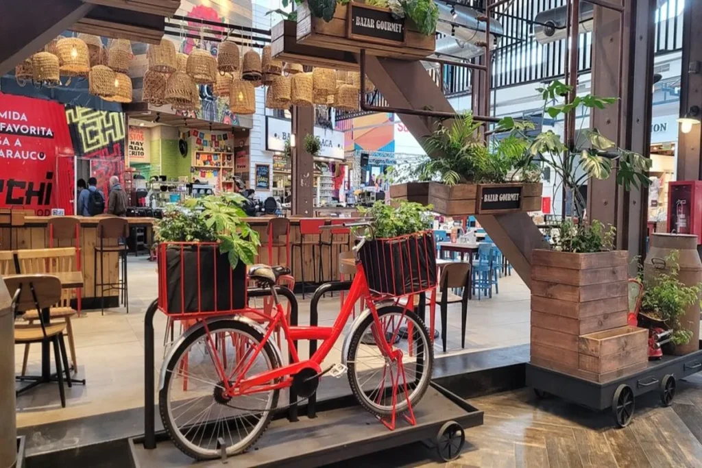 Interior de uma praça de alimentação com decoração rústica e industrial, com uma bicicleta vermelha decorativa, luminárias de vime e vegetação, dentro de um shopping center.