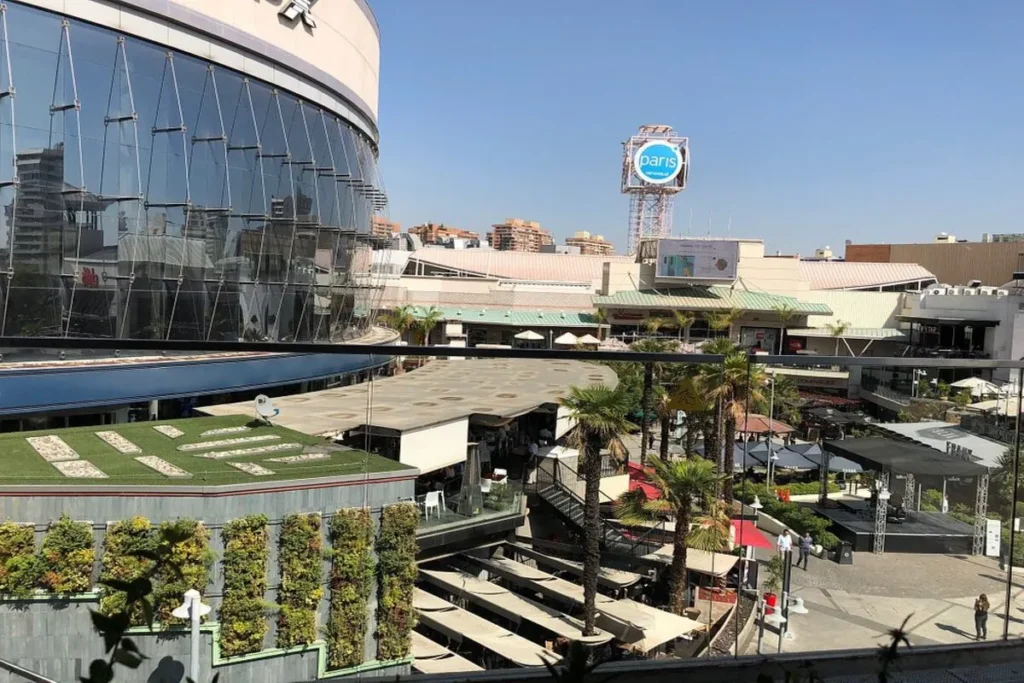 Vista aérea do shopping a céu aberto Parque Arauco em Santiago, com áreas de convivência, restaurantes, palmeiras e um edifício moderno com fachada de vidro.