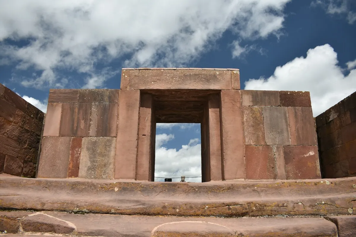 Vista de dentro para fora de uma porta de pedra monumental no sítio arqueológico de Tiwanaku, com céu azul e nuvens visíveis através da abertura.