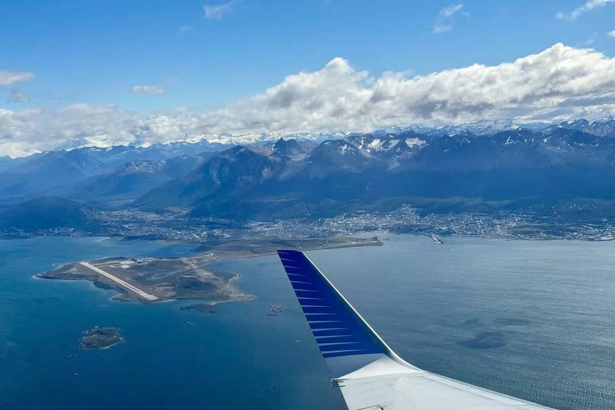 Vista aérea do Aeroporto Internacional de Ushuaia, da cidade e das montanhas nevadas, com a asa de um avião em primeiro plano.