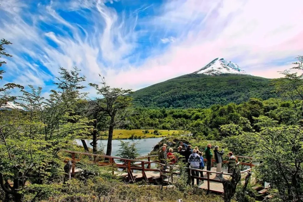 Grupo de turistas, incluindo um brasileiro em Ushuaia, em uma passarela de madeira com vista para um lago e montanhas nevadas.