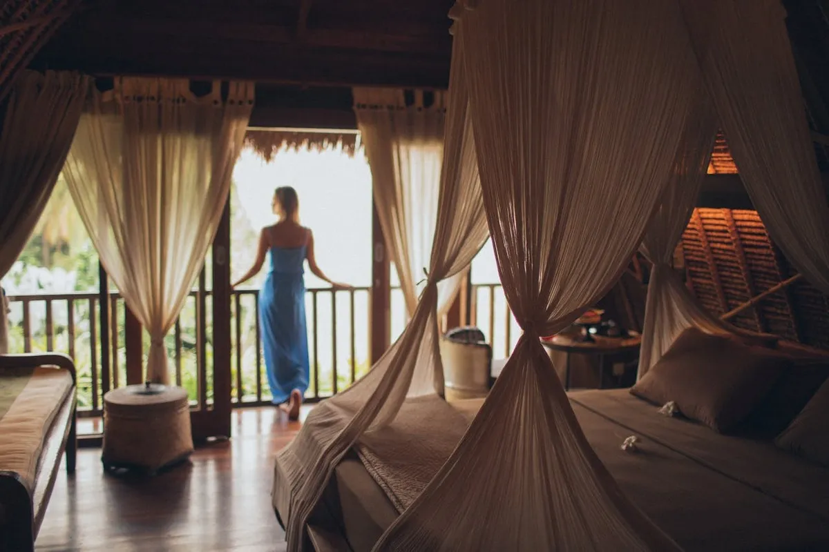 Interior de quarto de hotel com cama de dossel e cortinas leves. Uma pessoa de costas na varanda, apreciando a vista.
