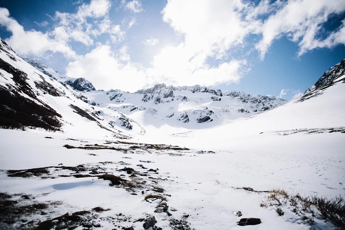 Vista das montanhas e vales cobertos de neve no Glaciar Martial, com céu azul e nuvens brancas iluminadas pelo sol.