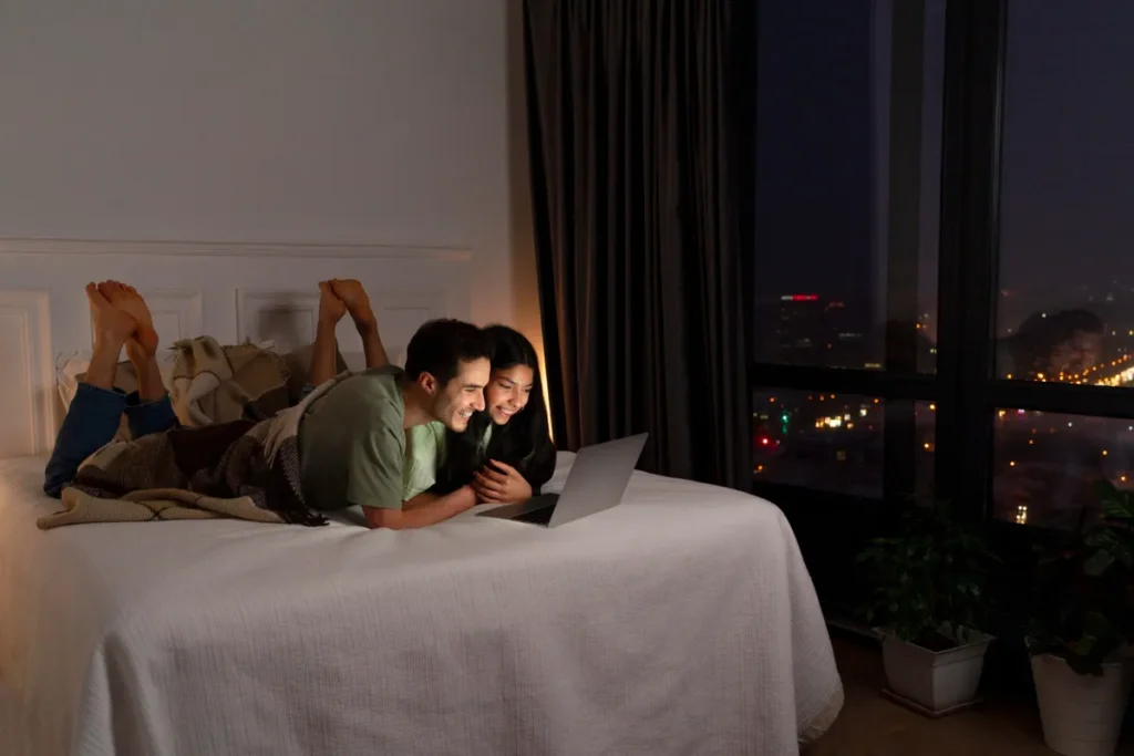 Casal assistindo a um filme em notebook na cama de um quarto de hotel à noite.