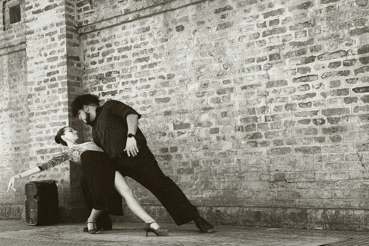 shows de tango em Buenos Aires, casal de dançarinos de tango em preto e branco em rua com parede de tijolos.